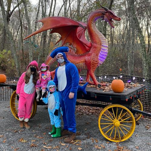 A family in colorful costumes poses by a dragon statue on a cart, surrounded by pumpkins in a wooded area.