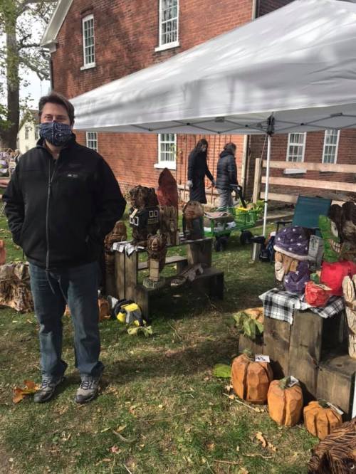 A man wearing a mask stands near a market stall displaying wooden sculptures and crafts under a tent.
