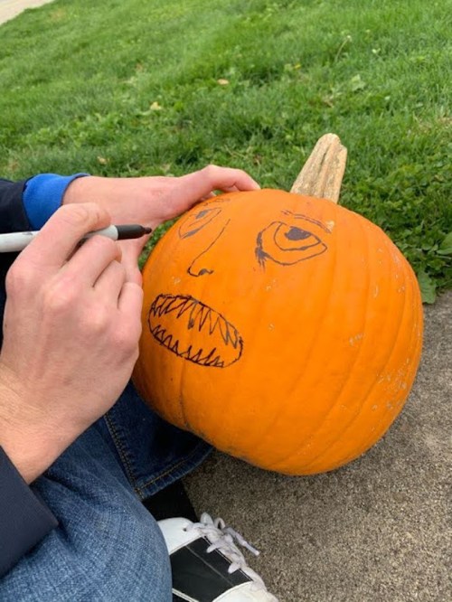 A person draws a face on a carved pumpkin sitting on the ground, surrounded by grass.
