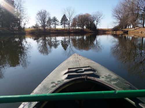 A kayak on calm water, reflecting trees and a clear blue sky, with a peaceful landscape in the background.