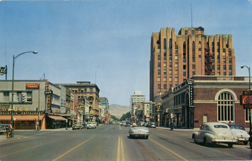 A vintage street scene featuring shops, restaurants, and classic cars, with a tall building in the background.