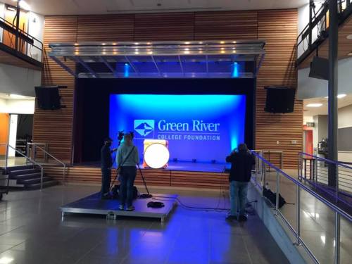 A stage setup at Green River College Foundation with blue lighting and a large logo backdrop. Two people are preparing equipment.