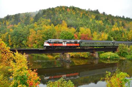 A colorful train crosses a bridge over a river, surrounded by vibrant autumn foliage and rolling hills.