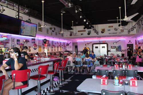 A retro diner interior with pink neon lights, red booths, and patrons enjoying food and drinks at the counter and tables.