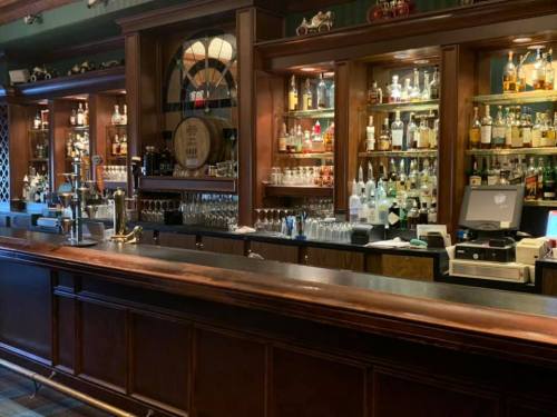 A wooden bar with shelves of various liquor bottles and glassware, illuminated by warm lighting.