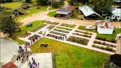 Aerial view of a rural area with gardens, a tractor, and a group of people walking along pathways near buildings.
