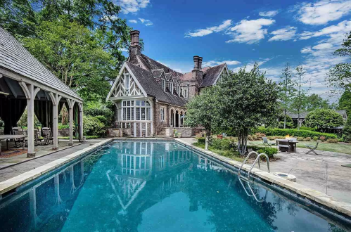 A luxurious home with a large pool surrounded by greenery and outdoor seating, under a bright blue sky.