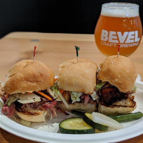 Three sliders with various toppings on a plate, accompanied by a glass of craft beer and pickles.