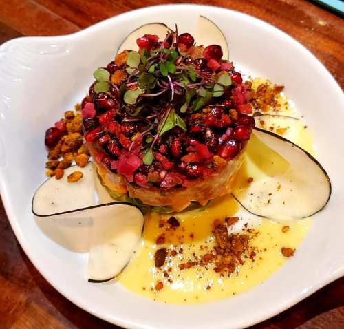 A colorful dish featuring layered fruits, pomegranate seeds, microgreens, and garnishes on a white plate.
