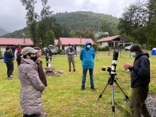 A group of people in outdoor clothing gathers around a telescope in a grassy area, with mountains in the background.