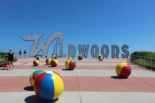 Colorful beach balls scattered on a walkway in front of a large "WILDWOODS" sign against a clear blue sky.