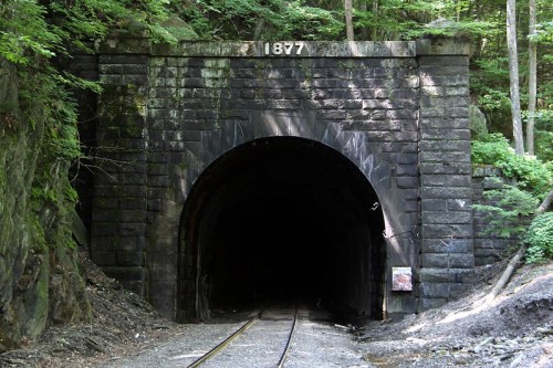 A dark tunnel entrance with a stone arch, marked "1877," surrounded by lush greenery and railway tracks.