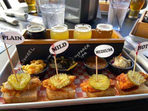 A tray with three fried chicken sliders topped with pickles, served with sides and a flight of craft beers.