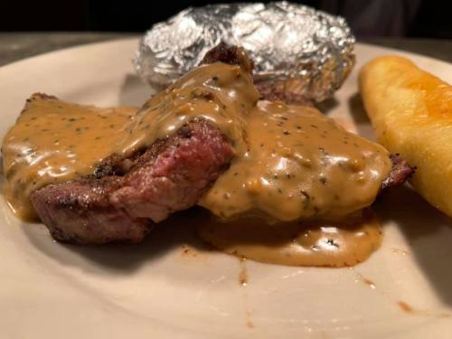 A plate featuring grilled steak topped with creamy sauce, a baked potato wrapped in foil, and a bread roll.