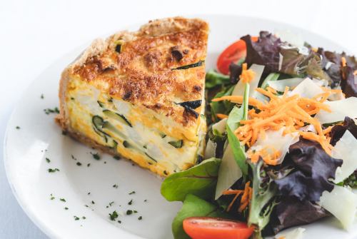 A slice of vegetable quiche served with a fresh salad of greens, tomatoes, and shredded carrots on a white plate.