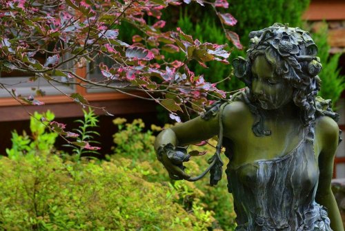 A green statue of a woman surrounded by foliage, holding a small bird, with a backdrop of trees and a wooden structure.