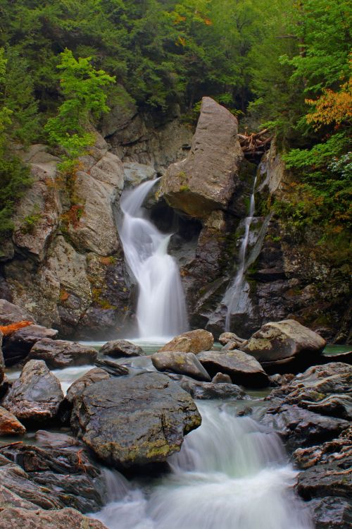 A serene waterfall cascades over rocky cliffs, surrounded by lush green trees and smooth stones in a tranquil forest setting.