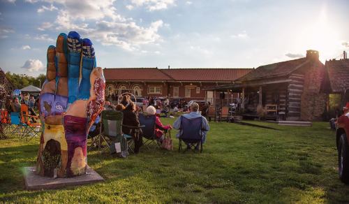 A colorful hand sculpture stands in a grassy area where people sit in chairs, enjoying an outdoor event near a log cabin.