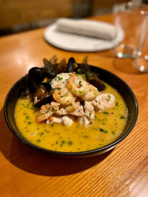 A bowl of seafood stew with shrimp, mussels, and herbs in a rich broth, served on a wooden table.