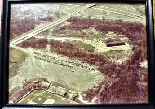 Aerial view of a vacant lot with a building, surrounded by trees and a highway, with parked vehicles in the distance.