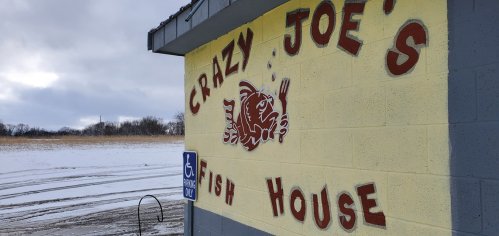 Sign for "Crazy Joe's Fish House" on a yellow wall, with a snowy landscape in the background.