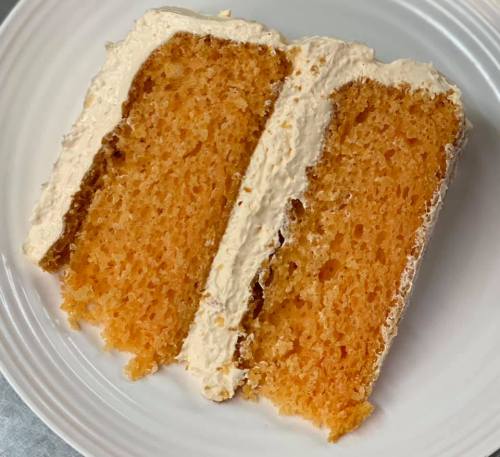 A slice of orange cake with creamy frosting on a white plate, showcasing its moist texture.