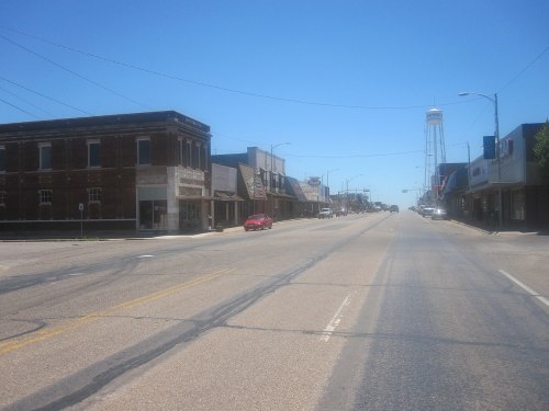 A quiet street scene featuring empty storefronts and a water tower under a clear blue sky.