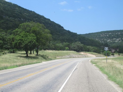 A winding road with a highway sign for Route 83, surrounded by green hills and trees under a clear blue sky.