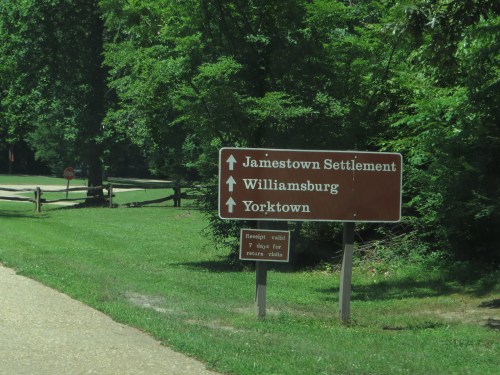 Brown directional sign indicating routes to Jamestown Settlement, Williamsburg, and Yorktown, surrounded by greenery.