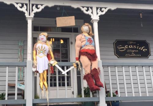 Two scarecrows dressed as superheroes hang from a porch, with a sign above them at a seaside spa salon.