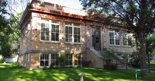 Historic brick building with large windows, surrounded by green grass and trees. Steps lead to the entrance.