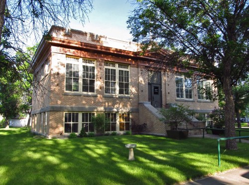 A brick building with large windows, surrounded by green grass and trees, featuring a set of stairs at the entrance.