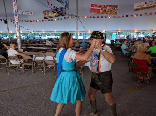 A woman in a blue dress dances with an older man in traditional attire under a festive tent filled with chairs.