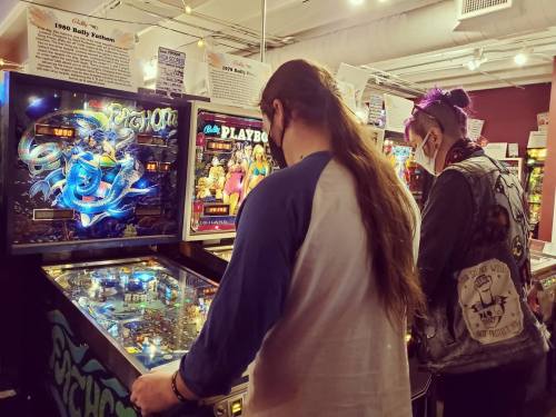Two people playing pinball at an arcade, focused on the machines with colorful graphics and retro designs.