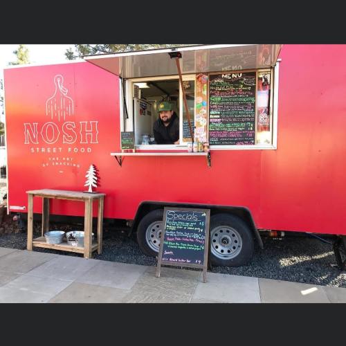 A food truck named "Nosh" with a menu displayed, featuring a vendor smiling from the window.