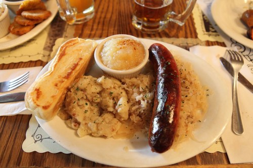 A plate of food featuring sausage, sauerkraut, mashed potatoes, and a side of applesauce, with a toasted bun.