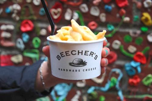 A hand holding a cup of mac and cheese from Beecher's Handmade Cheese against a colorful wall.