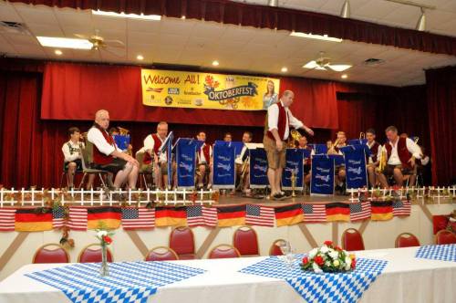 A band in traditional attire performs on stage at an Oktoberfest event, with flags and decorations in the background.