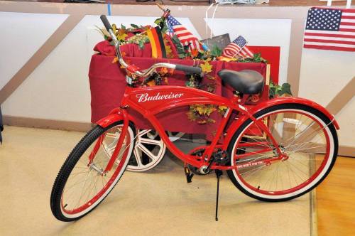 A red Budweiser bicycle is displayed next to a decorated table with flags and greenery.