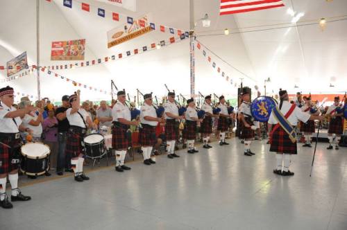 A group of bagpipers in traditional attire perform indoors, while an audience claps and enjoys the music.
