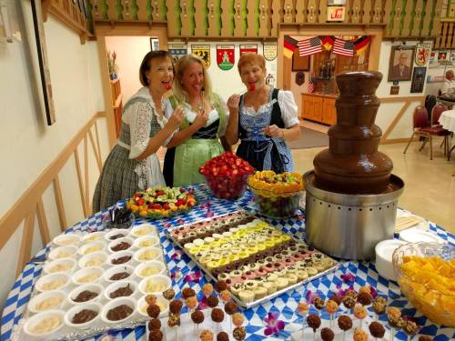 Three women in traditional dresses pose by a dessert table featuring a chocolate fountain and various treats.