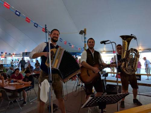 A lively band performs under a tent, featuring an accordion, guitar, and tuba, with an audience enjoying the music.