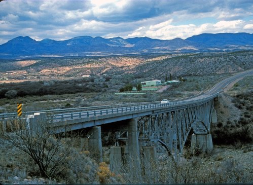 A bridge spans a valley with mountains in the background under a cloudy sky, showcasing a scenic landscape.