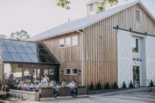 A rustic wooden barn with a glass extension, featuring outdoor dining and guests enjoying a meal.
