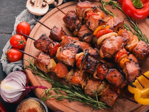 Grilled meat skewers with vegetables on a wooden platter, surrounded by fresh herbs and spices.