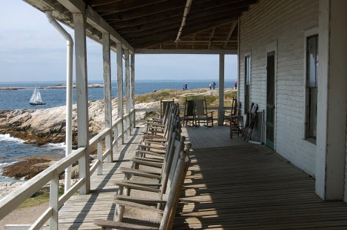 A seaside porch with rocking chairs overlooks the ocean, with a sailboat in the distance and people walking along the shore.