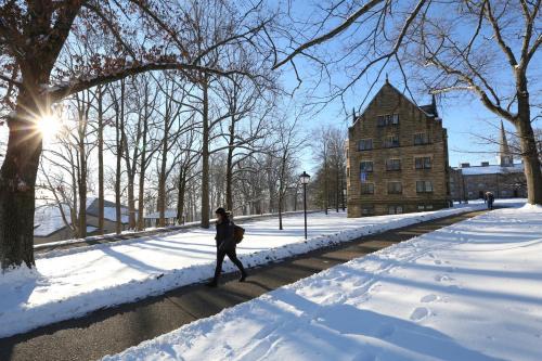 A person walks along a snowy path beside a historic building, with trees and sunlight in the background.