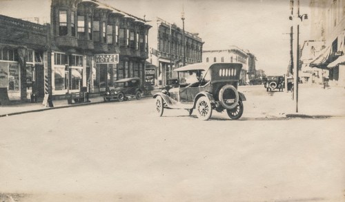Black and white photo of a vintage car on a street with historic buildings and a hotel in the background.