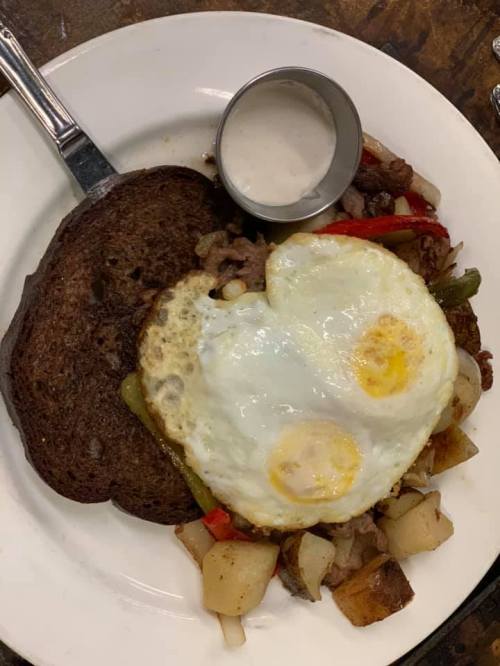 A plate with two sunny-side-up eggs, sautéed vegetables, potatoes, and a slice of dark bread, served with a small cup of sauce.