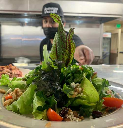 A vibrant salad with greens, tomatoes, and toppings, with a chef in the background reaching out.
